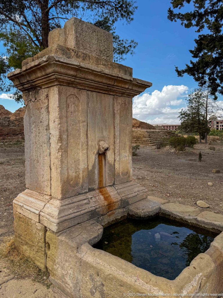 Rectangular stone fountain basin below a masonry fountain structure with a projecting spout and water staining at Timgad.