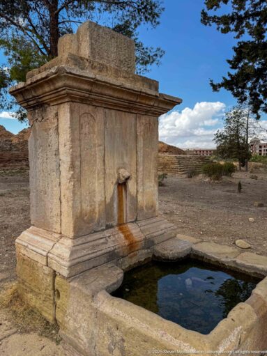 Rectangular stone fountain basin below a masonry fountain structure with a projecting spout and water staining at Timgad.