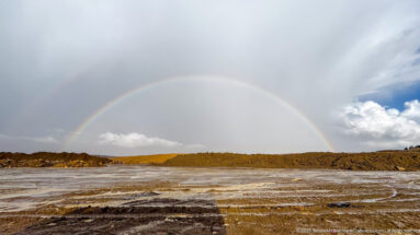 Nearly complete rainbow spanning gray sky above pale open ground and a low ridge near Timgad.