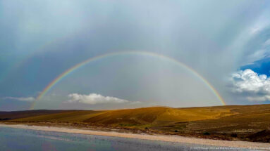 Primary and faint secondary rainbow arching over rolling golden fields beneath dark storm clouds near Timgad.