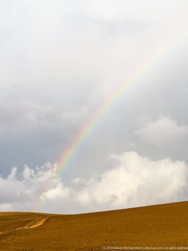 Diagonal rainbow descending toward golden-brown fields beneath layered gray clouds near Timgad.