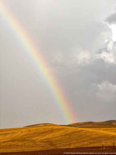 Close view of a bright rainbow descending toward ochre fields beneath gray rain clouds near Timgad.