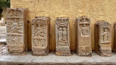 Five carved stone stelae with human figures and relief ornament displayed upright against an ochre wall.