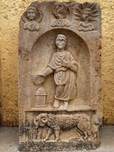 Stone stele with a standing robed figure in a niche, upper symbols, and an animal relief along the base.