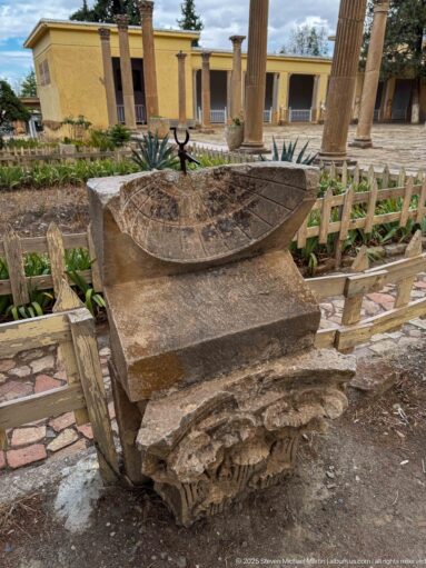 Carved stone sundial with incised radial hour lines displayed on a block in a courtyard of ancient fragments.