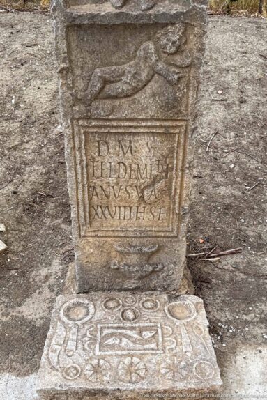 Weathered stone monument with a Latin inscription, reclining animal relief above, and geometric decoration below.