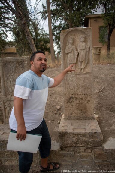 Guide standing beside a carved stone monument and pointing to its relief during an explanation at Timgad.