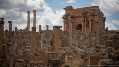 Large Roman arched monument with engaged columns rising above scattered columns and masonry ruins at Timgad.