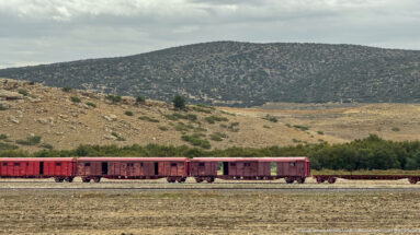 Freight train crossing a dry plain below a low scrub-covered hill near Timgad.