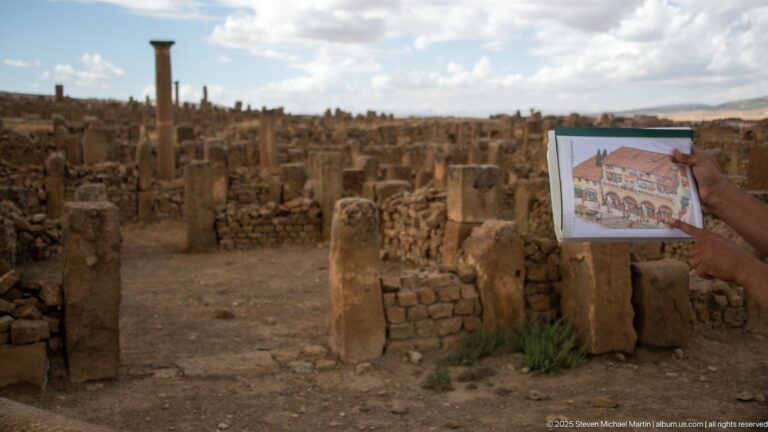 Hand holding a color reconstruction image in front of the corresponding masonry ruins at Timgad.