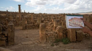 Hand holding a color reconstruction image in front of the corresponding masonry ruins at Timgad.
