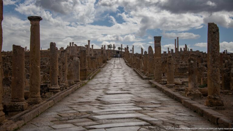 Long paved Roman street lined by columns and ruins beneath dramatic clouds at Timgad.