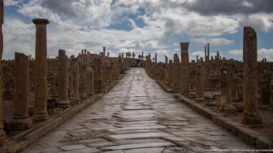 Long paved Roman street lined by columns and ruins beneath dramatic clouds at Timgad.
