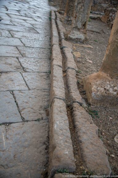 Close view of large street paving slabs beside a raised stone curb and narrow drainage channel.