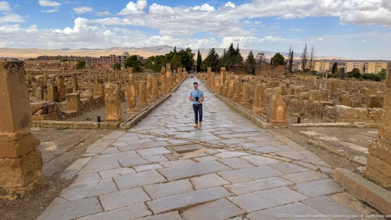 Visitor walking down a broad stone-paved Roman street lined with columns and ruins at Timgad.