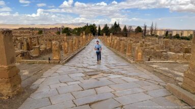 Visitor walking down a broad stone-paved Roman street lined with columns and ruins at Timgad.