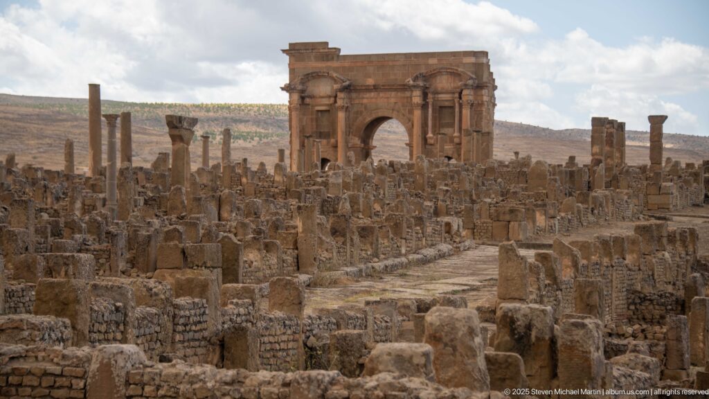 Arch of Trajan rising beyond dense Roman ruins and a paved street corridor at Timgad.