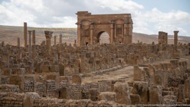 Arch of Trajan rising beyond dense Roman ruins and a paved street corridor at Timgad.