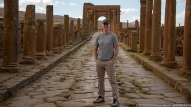 Visitor standing on Timgad’s paved colonnaded street with the Arch of Trajan behind him.