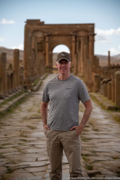 Visitor posing on an ancient paved street with the Arch of Trajan blurred in the background.