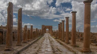 Symmetrical view down a paved colonnaded street toward the Arch of Trajan under dramatic clouds.