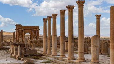 Tall Roman columns with ornate capitals and the Arch of Trajan visible beyond at Timgad.