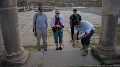 Four visitors gathered between large Roman columns as one bends toward the ancient pavement.