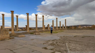 Tall Roman columns bordering a broad paved precinct with a lone visitor beneath dramatic clouds.