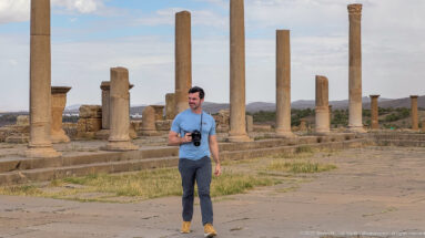 Visitor holding a camera on ancient paving among tall standing Roman columns at Timgad.