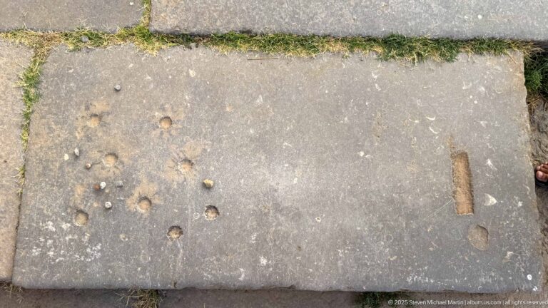 Large stone paving slab with scattered circular depressions and elongated cut marks beside a curb.