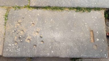Large stone paving slab with scattered circular depressions and elongated cut marks beside a curb.
