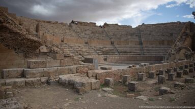 Semicircular stone seating and stage remains inside the Roman theater at Timgad, with masonry blocks in the foreground.
