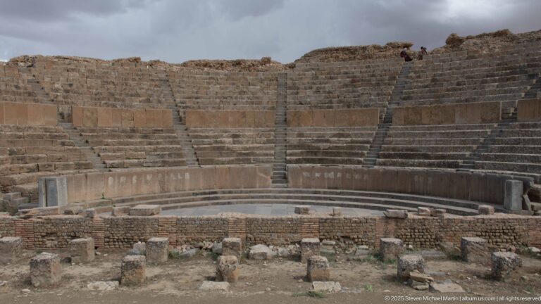 Centered view across the orchestra toward curved tiers of stone seating in Timgad’s Roman theater.
