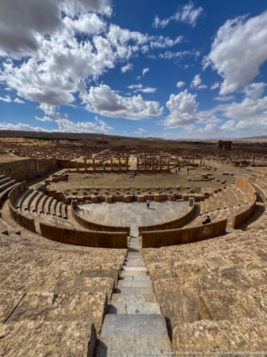 High central view over Timgad’s Roman theater toward the archaeological city and distant plateau.