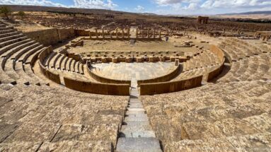 Wide elevated view of the Roman theater at Timgad, showing curved seating, paved orchestra, and surrounding ruins.