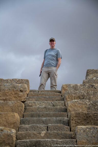 Visitor standing above a steep stone stairway between seating blocks in Timgad’s Roman theater.