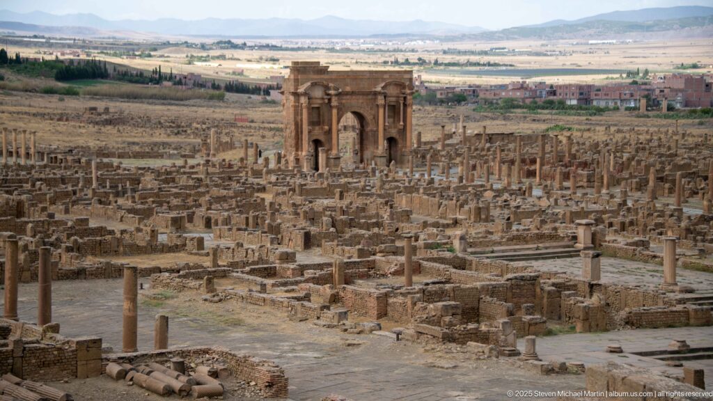 Arch of Trajan rising above the extensive Roman ruins of Timgad, with modern buildings and hills beyond.