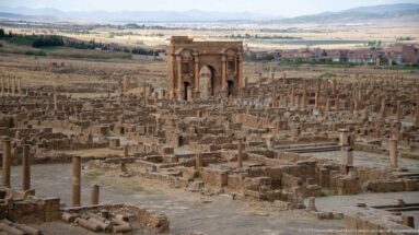 Arch of Trajan rising above the extensive Roman ruins of Timgad, with modern buildings and hills beyond.