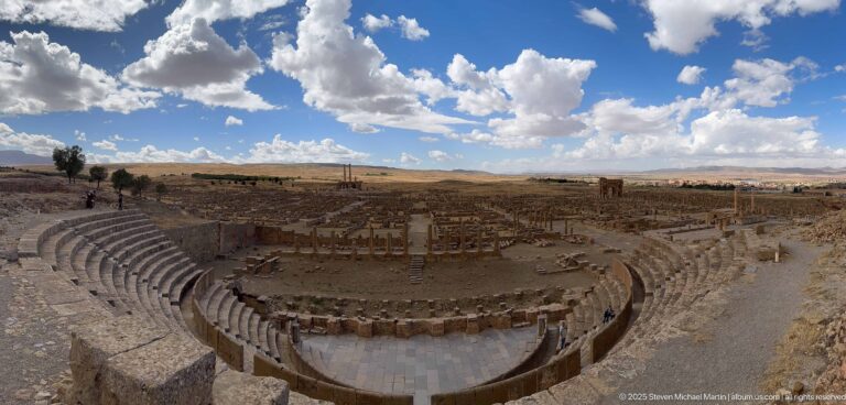 Ultra-wide view of Timgad’s Roman theater, archaeological city, open plateau, and cloud-filled sky.