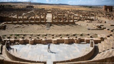 Steep view from the upper cavea down to the paved orchestra and stage remains of Timgad’s Roman theater.