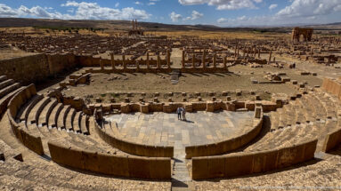 Elevated centered view of Timgad’s theater with paved orchestra, curved seating, stage remains, and city ruins beyond.