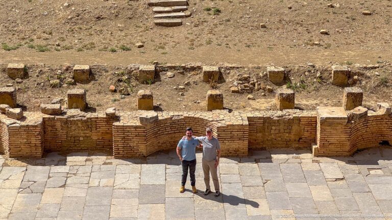 Two visitors standing on the paved orchestra of Timgad’s Roman theater below low stage remains.