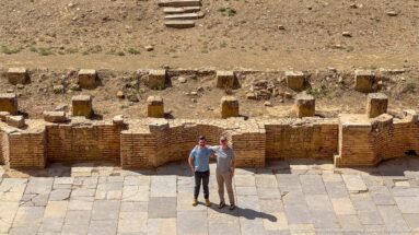 Two visitors standing on the paved orchestra of Timgad’s Roman theater below low stage remains.