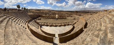Wide-angle view inside Timgad’s Roman theater with steep curved seating around the paved orchestra.