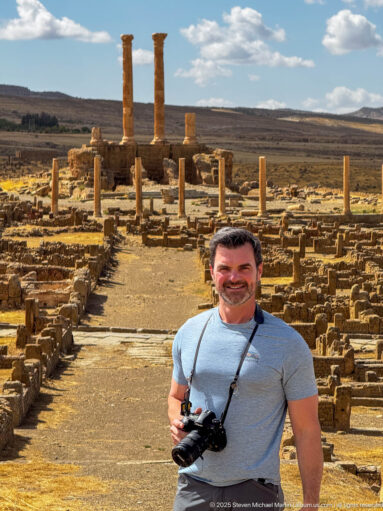Visitor holding a camera in front of Timgad’s ruins, with standing columns aligned along an ancient street behind him.