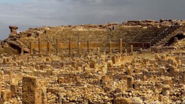 Dense stone foundations and columns at Timgad with the high curved seating of the Roman theater behind.