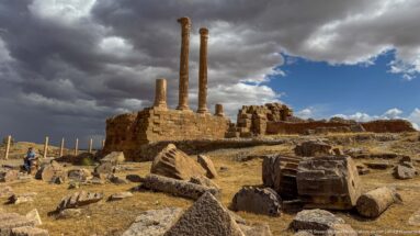 Two tall Roman columns rising above ruined masonry and scattered architectural blocks under dark storm clouds at Timgad.