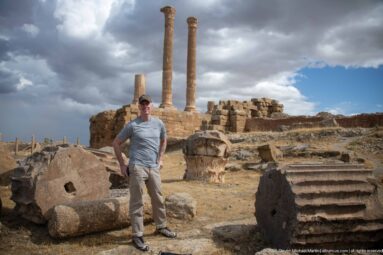 Visitor standing among large carved capitals, column fragments, and monumental Roman ruins at Timgad.