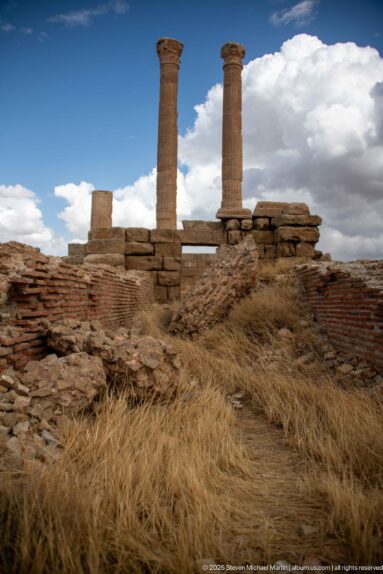 Two tall Roman columns rising above layered brick and stone ruins with dry grass in the foreground at Timgad.