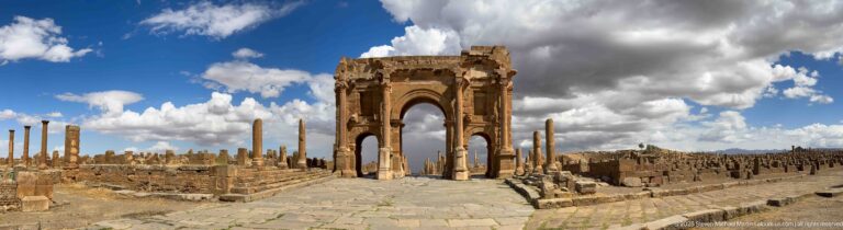 Tall single-bay Roman arch centered on a paved ancient route, with columns and low ruins extending on both sides at Timgad.